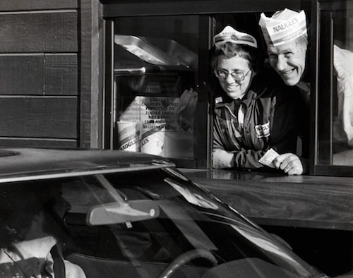 Two people wearing Naugles fast food hats smile outside of a drive-thru window.