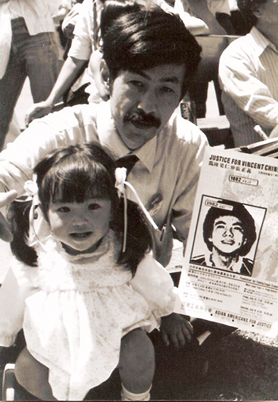 A man with his child hold up a flyer that reads Justice for Vincent Chin.